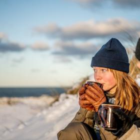 Jente holder varmen med en kopp varm drikke ved Klitmøller strand om vinteren